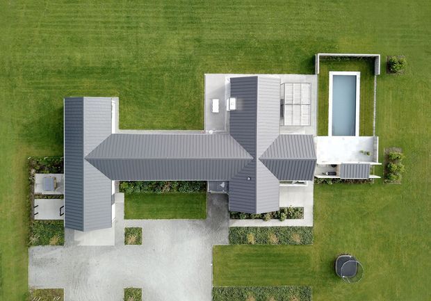 Aerial top-down view of a large contemporary residential property set within expansive manicured lawns. The building has several interconnected wings with dark grey pitched metal roofs, light-coloured exterior walls, paved circulation areas, landscaped garden strips, and a rectangular swimming pool beside a covered outdoor terrace.