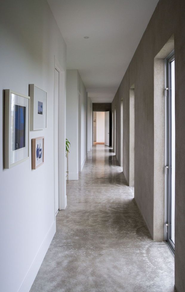 A long, narrow minimalist hallway with a polished grey concrete floor, white walls and ceiling, and a dark textured wall containing a series of tall recessed windows. Framed artwork and a small plant line the opposite wall, while open doorways lead to rooms at the far end.