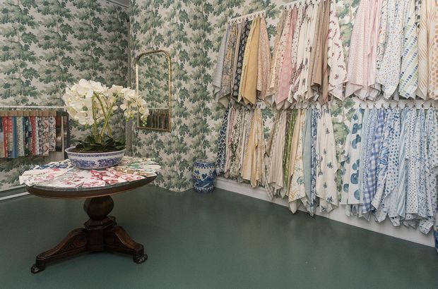 A richly patterned textile showroom or fabric display room, with numerous hanging fabric samples in floral, paisley, and geometric prints arranged along the walls. The room features botanical wallpaper, a dark green painted lower wall and floor, a round antique-style wooden pedestal table, a patterned ceramic planter with white flowers, and a small wall mirror.