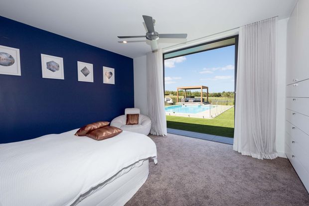 A bright, contemporary bedroom with a large white bed, deep navy feature wall, framed artwork, white built-in drawers, and pale carpet. Full-height sheer curtains open onto a landscaped pool area visible through expansive glazing, while a ceiling fan provides ventilation. The room has a clean, relaxed and resort-like atmosphere.