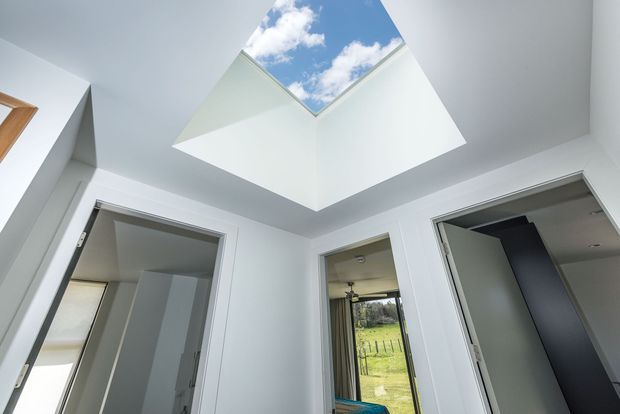 Bright white interior landing or hallway viewed upward toward a large square skylight, with blue sky and clouds visible above. Several open doorways connect to adjacent rooms, including a glimpse of a green rural landscape through a glazed opening. The space has clean white walls, simple white door frames, and a contemporary minimalist architectural character.
