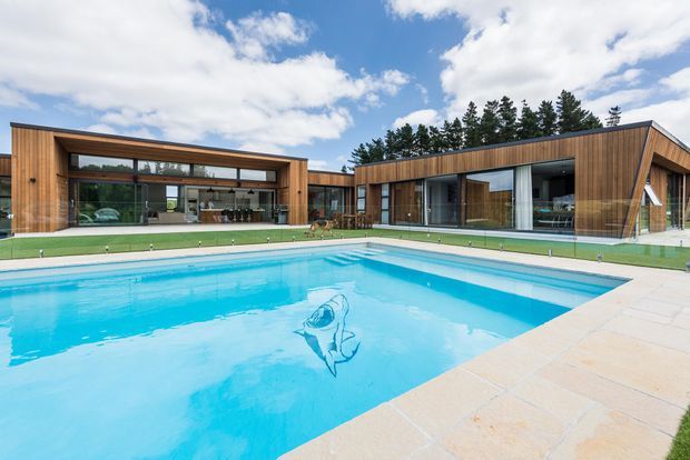 A low, expansive modern residence arranged around a large swimming pool. The home features strong horizontal lines, extensive floor-to-ceiling glazing, warm timber-clad exterior walls, dark metal framing, and flat or very low-pitched roof forms. A pale paved pool terrace and manicured lawn sit in the foreground, with a wooded backdrop under a partly cloudy sky.