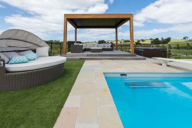 A landscaped outdoor pool area with a bright blue swimming pool bordered by pale stone paving. A raised timber-framed pavilion contains an outdoor lounge with grey sofas, while a rounded woven daybed sits on artificial grass in the foreground. Open rural fields and a partly cloudy sky are visible beyond the property.
