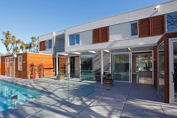 A contemporary two-storey residential home viewed from its pool terrace. The building has a crisp white rendered facade, extensive glazing, warm timber louvred shutters and timber-clad sections, with a glass pool fence, paved terrace and swimming pool in the foreground. Palm-like planting and strong sunlight create an open, resort-like outdoor setting.