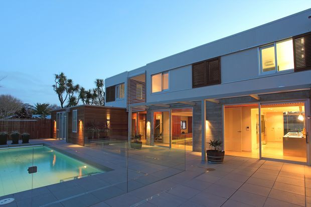 Twilight exterior view of a contemporary two-storey residence with a flat roof, large glazed ground-floor openings, timber privacy screens, and a paved terrace surrounding a swimming pool. Warm interior lighting contrasts with the cool blue-grey facade and evening sky.