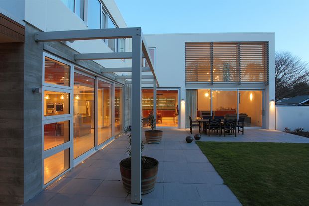 Modern two-storey residential courtyard exterior at dusk, featuring extensive floor-to-ceiling glazing, a rendered white facade, exposed concrete elements, timber horizontal sun louvres, and a paved outdoor dining terrace with lawn. Warm interior lighting glows through the glass doors and windows.