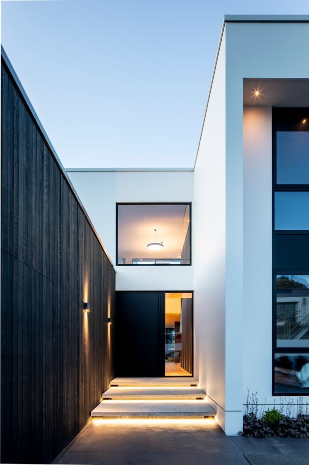 A dusk exterior view of a narrow contemporary residential entry courtyard. Illuminated concrete steps lead to a black pivot-style front door, flanked by a tall dark vertical timber privacy fence on the left and a crisp white two-storey facade on the right. Large black-framed windows reveal warm interior lighting, while recessed and wall-mounted lighting accentuates the entrance and stair edges.