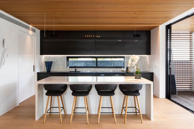 Contemporary kitchen with a large white island and four black bar stools, dark full-height cabinetry, a black benchtop, integrated horizontal window, and warm timber ceiling lining. White marble-look splashback panels and light timber flooring create a bright, refined interior. Timber screening and an open glazed doorway are visible at the right.