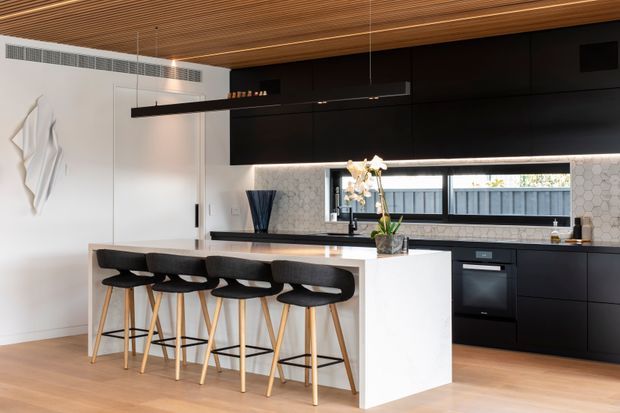 Contemporary open-plan kitchen with a large white island and four black upholstered bar stools, matte black full-height cabinetry, integrated appliances, a pale hexagonal tile splashback, and a horizontal window above the benchtop. The room features warm timber flooring and a slatted timber ceiling with slim linear pendant lights, creating a refined, high-contrast interior.