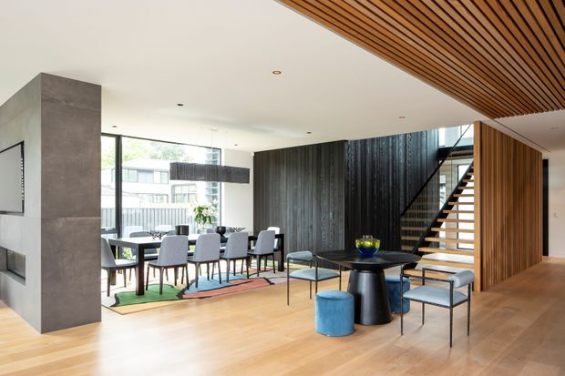 Open-plan contemporary dining and living area with a large dining table, sculptural coffee table and stools, expansive glazing, a dramatic dark timber feature wall, and a staircase framed by warm vertical timber battens. The room has light timber flooring, a white ceiling, and a striking slatted timber ceiling detail.
