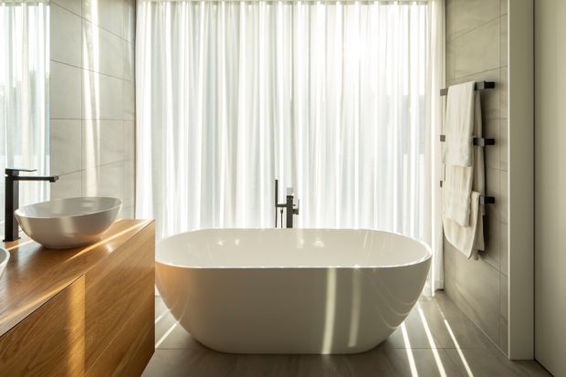 Bright contemporary bathroom featuring a freestanding white oval bathtub, a timber vanity with a vessel basin, dark minimalist tapware, large pale wall tiles, and full-height sheer curtains filtering abundant natural light. A wall-mounted towel rail with folded towels is visible on the right.