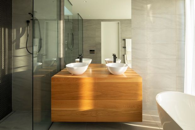 Contemporary bathroom with a large floating timber double vanity, twin white vessel basins and black tapware. A frameless glass shower enclosure occupies the left side, while large pale stone-look wall tiles surround the room. A freestanding white bathtub is partially visible at right, with a full-width mirror reflecting the vanity and shower. Strong natural light creates a warm, refined atmosphere.