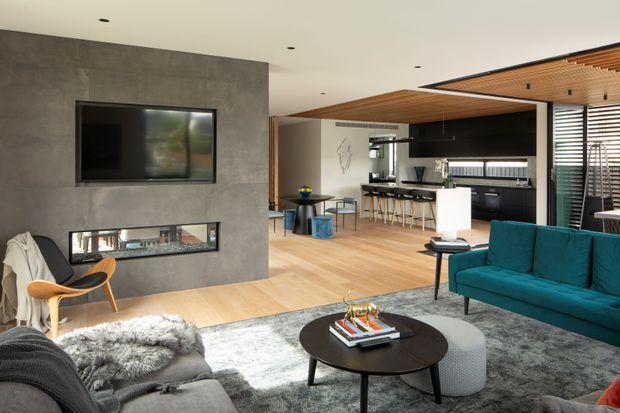 Open-plan contemporary living area with a large grey feature wall containing a wall-mounted television and linear fireplace. The room includes a teal sofa, grey sectional seating, round black coffee table, textured rug, timber flooring, and an adjacent kitchen with a dark island and bar stools. A timber-lined ceiling, black-framed glazing, and horizontal privacy blinds contribute to the modern interior.
