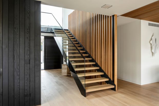 A refined contemporary entry hall dominated by an open-riser timber staircase with slim black metal stringers and a clear glass balustrade. A full-height vertical timber slat screen forms a feature wall beside the stairs, while dark vertical timber wall panelling frames the left side. Light timber flooring, a white plaster wall, recessed ceiling lighting, and a timber slatted ceiling detail create a warm, minimalist interior.