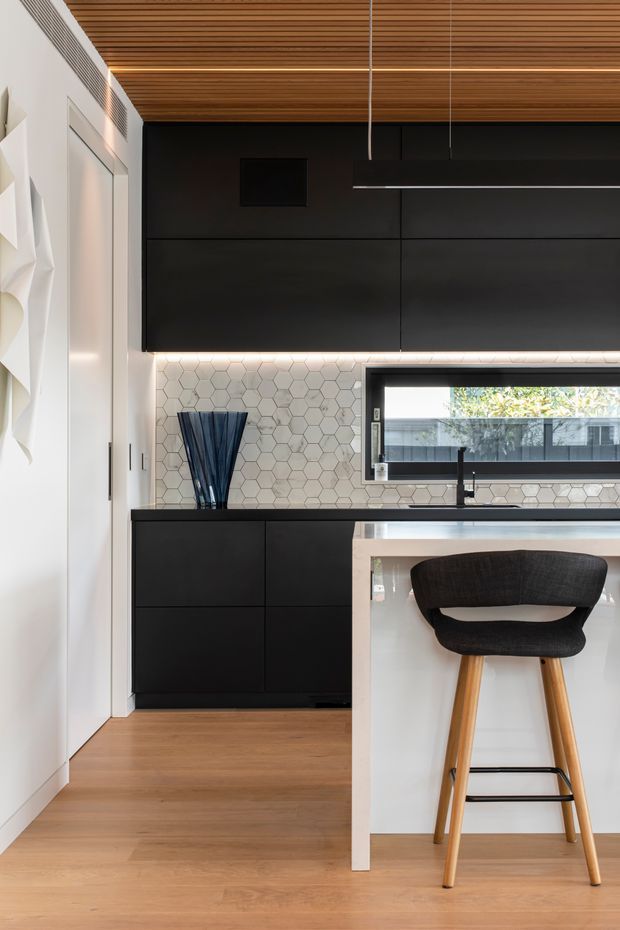 A compact contemporary kitchen with matte black full-height cabinetry, a white island with a dark benchtop, and a slim black-framed horizontal window. The splashback is clad in small light hexagonal tiles, while the ceiling features warm timber slats with integrated linear lighting. Light timber flooring, a black pendant light, and a black-and-timber bar stool complete the refined monochromatic scheme.