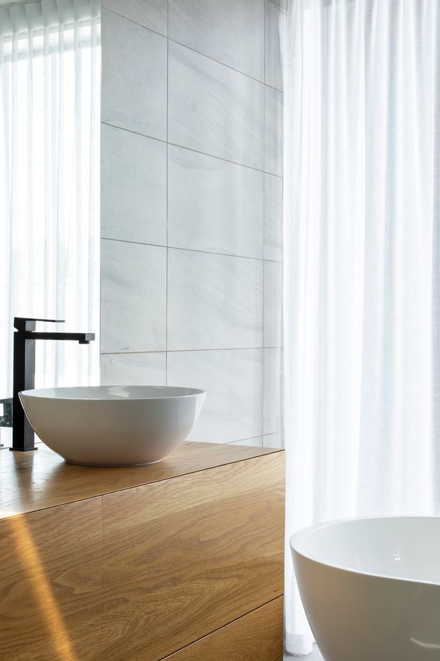 Bright, refined contemporary bathroom featuring a freestanding white vessel basin on a warm timber vanity, a matte black wall-mounted tap, large pale marble-look wall tiles, sheer white curtains, and part of a freestanding bathtub in the foreground.