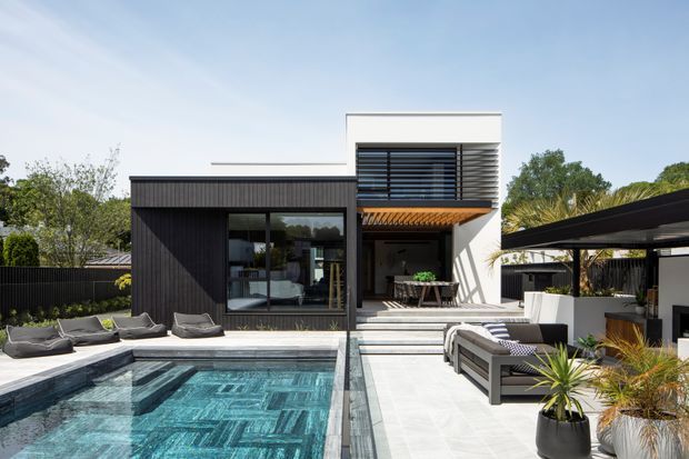 Contemporary two-storey residence with a dark vertically clad lower level and crisp white upper volume. Broad glazing, horizontal louvres, timber-toned canopy and a covered outdoor entertaining area open onto a pale-paved terrace and swimming pool. The setting includes landscaped planting and a private courtyard-like backyard.