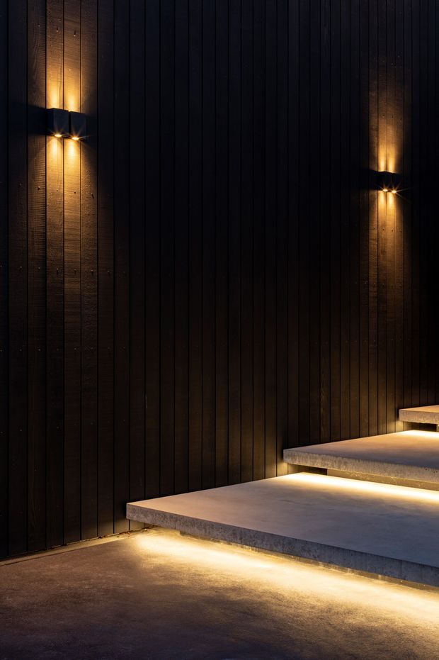 A dark, dramatic architectural detail showing broad concrete steps floating against a tall black vertically clad wall. Warm recessed under-step lighting and wall-mounted sconces create a strong contrast, highlighting the textured concrete and timber-like vertical facade.