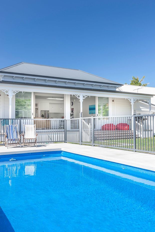 Bright exterior view of a white heritage-style single-storey home with a broad covered verandah, decorative fretwork, white railings and a corrugated metal hipped roof. A vivid blue swimming pool and pale paved pool deck dominate the foreground, with glimpses of the open-plan kitchen and living area behind the verandah.