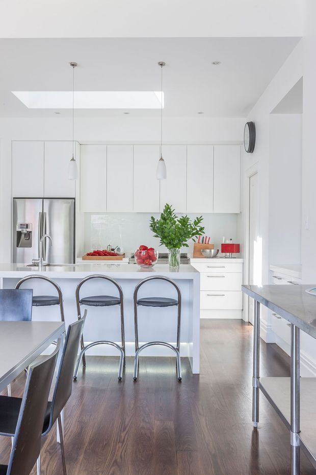 Bright, predominantly white contemporary kitchen viewed from the dining area. The room features a central island with seating, full-height white cabinetry, stainless-steel refrigerator and appliances, a pale reflective backsplash, pendant lights, skylight, and warm timber flooring. A dining table and chairs occupy the foreground, while a stainless-steel-topped work surface is visible at right.