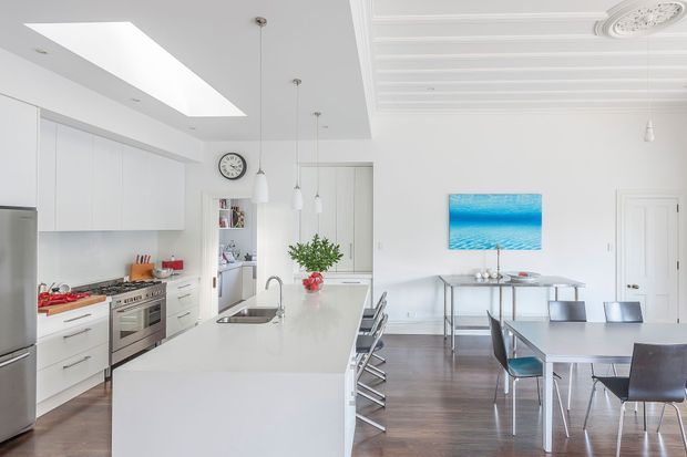 Bright, open-plan kitchen and dining area with a large white island, integrated white cabinetry, stainless-steel appliances, pendant lights, and a dining table with contemporary chairs. The room features a high decorative ceiling with ornate ceiling medallions and moulding, a large skylight, timber flooring, and a vivid blue artwork on the wall.