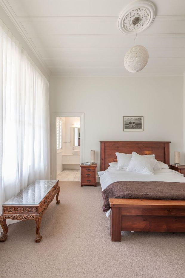 A bright traditional bedroom with a large dark timber bed, white bedding, a brown throw, matching timber bedside tables, and an ornate carved timber coffee table. Floor-to-ceiling sheer curtains line the left wall, while decorative ceiling mouldings and a ceiling medallion surround a woven pendant light. A bathroom is visible through the open doorway at the rear.