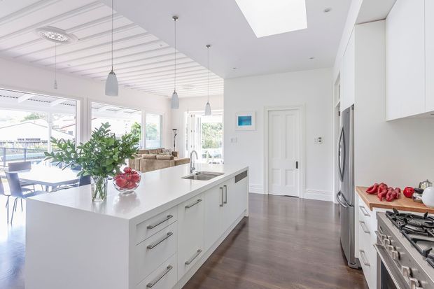 Bright, spacious white kitchen with a large central island, integrated storage, stainless-steel appliances, timber-look flooring and extensive glazing. The island opens toward a dining and living area, with a coffered ceiling, pendant lights and a large skylight adding architectural detail. The room has a clean, refined and welcoming appearance.