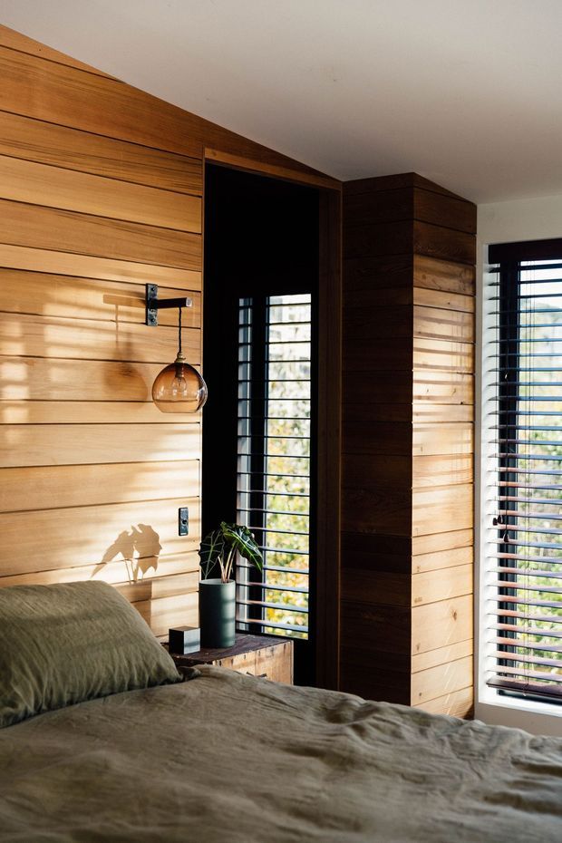 A warmly lit bedroom corner with extensive horizontal timber wall panelling, a dark timber-framed window and structural column, black louvred blinds, and a low bed dressed in muted olive-grey bedding. A small pendant light, potted plant, and bedside furniture add understated natural character. Strong sunlight creates striped shadows across the timber surfaces.