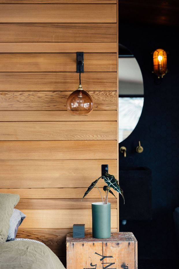 Close-up interior view of a warm timber-lined bedroom wall with horizontal wood boards, a hanging amber glass pendant light, and a rustic timber bedside table with a teal vase and plant. Part of a bed is visible at lower left. A dark adjoining space or doorway with a round mirror and warm wall light appears on the right.