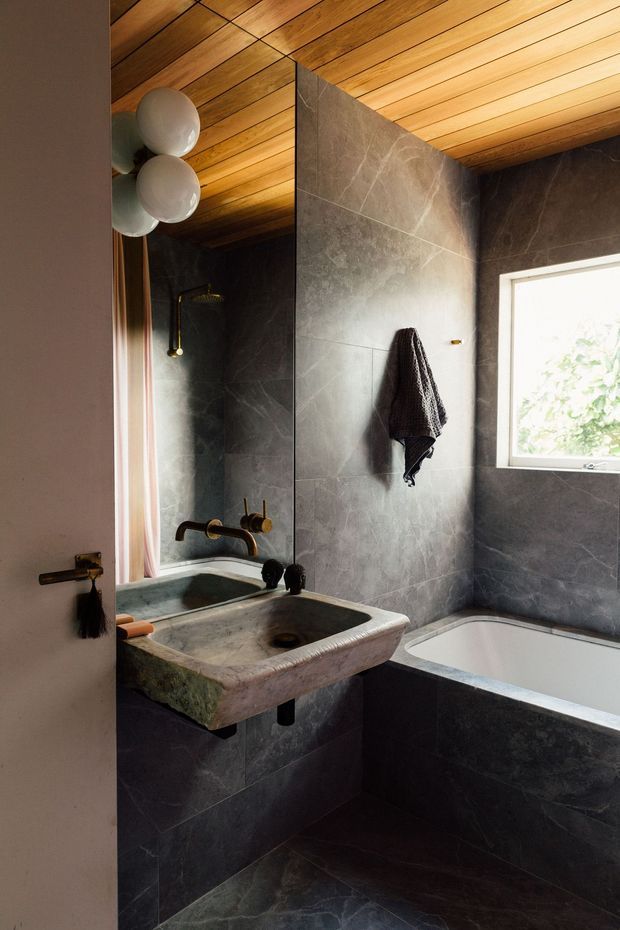 A compact, moody bathroom with dark stone-look wall and floor tiles, a carved natural stone wall-mounted basin, brass-toned tapware and shower fittings, and a built-in bathtub beneath a bright window. The ceiling is clad in warm timber boards, with globe wall lights and a large mirror adding warmth and depth.
