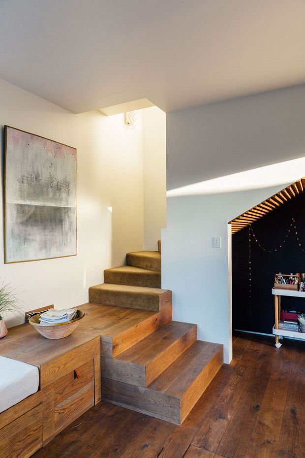 Interior view of a warm contemporary home featuring a compact timber staircase with integrated built-in storage and a carpeted upper stair run. White plaster walls and ceiling are illuminated by strong natural light, while a dark recessed nook with exposed timber slats and shelving is visible to the right. A framed abstract artwork and small furnishings add residential character.