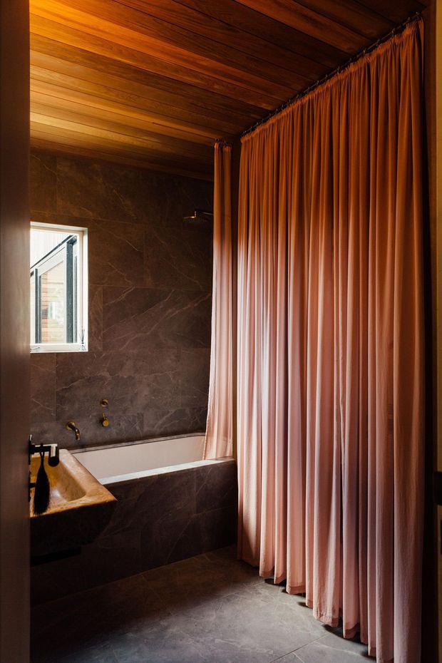 A moody, intimate bathroom featuring a freestanding-style bathtub set against dark marble-look wall and floor finishes, warm timber ceiling boards, brass tapware, and a full-height pleated blush-pink curtain enclosing the bathing area. A small window provides natural light.