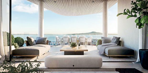 A bright, open-plan coastal living area with a large curved or gently projecting terrace overlooking the water. The room features pale upholstered seating, a low rectangular coffee table, a large rounded ottoman, white cylindrical columns, expansive glazing, and a white slatted ceiling. An outdoor dining area is visible beyond the living zone, with a calm bay and distant landforms in the background.