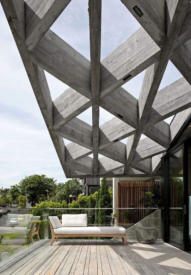 A modern elevated outdoor terrace viewed from below a dramatic exposed concrete canopy with large intersecting beams and integrated recessed lights. The terrace has pale timber decking, a frameless glass balustrade, contemporary outdoor seating, and a black-framed glazed wall beside a vertical timber privacy screen. Suburban greenery and a street are visible beyond the railing.