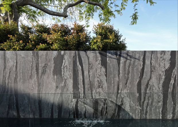 Exterior architectural detail showing a dark grey stone-clad water feature or pool wall beneath mature trees and planting. Large vertically veined stone panels form the main facade, with water spilling from a narrow rectangular outlet into a dark pool or basin. Strong sunlight and tree shadows create a dramatic, natural setting.