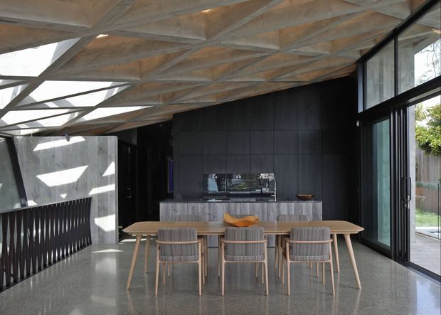 A contemporary dining area with a long light-wood dining table and matching chairs, positioned in front of a dark kitchen island and black tiled feature wall. The room has polished concrete flooring, extensive black-framed glazing, and an exposed geometric timber roof structure with strong natural light.