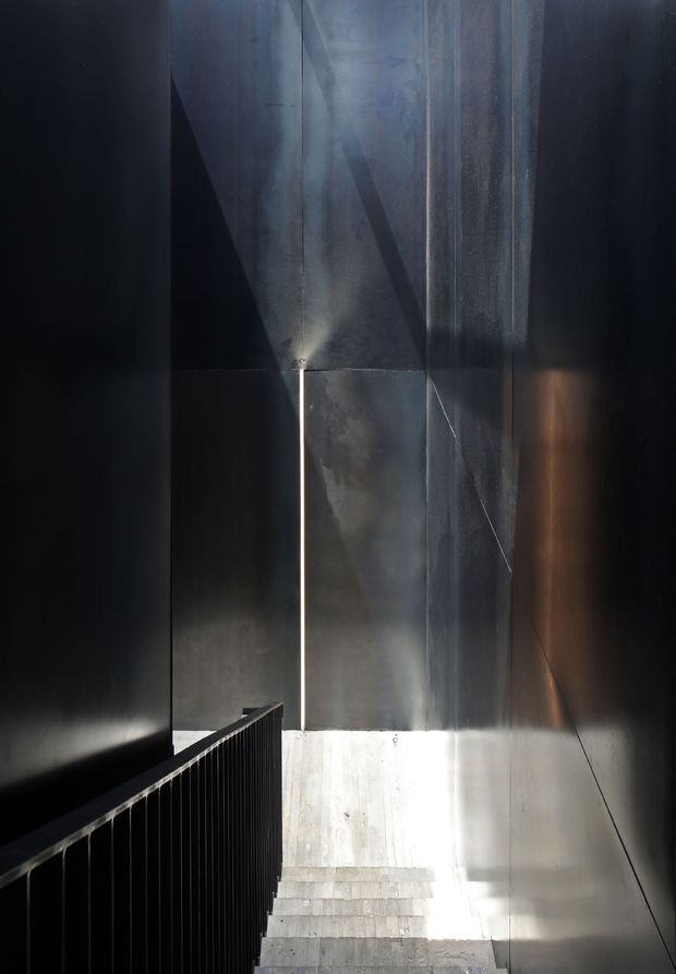 A dramatic, narrow stairway descending between tall dark metal-clad walls. A black vertical-slat balustrade lines the left side, while strong natural light enters from below and creates sharp highlights and shadows across the reflective wall surfaces and concrete steps. The composition is minimalist, moody, and architectural.