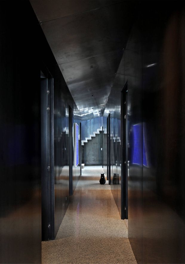 A dark, dramatic interior hallway with glossy black wall and ceiling panels, multiple doorways, reflective surfaces, and a polished terrazzo-like floor. Blue accent lighting glows from wall features, while a concrete stairway is visible at the far end, creating a striking contemporary circulation space.