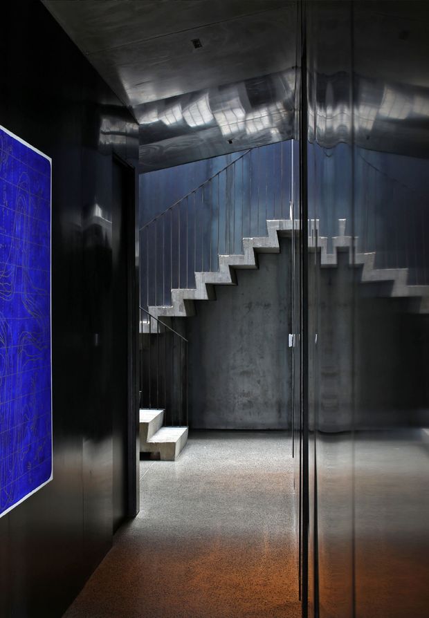 Dark, dramatic interior corridor focused on a sculptural concrete stair. The space features polished dark metal wall and ceiling surfaces, reflective glazing or mirrored panels, a raw concrete stair and wall, and a speckled polished floor. A vivid blue graphic artwork is partially visible on the left wall. The stair rises along the rear wall with a simple slender metal balustrade.