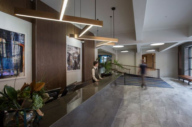 A refined commercial lobby or reception area with a long dark stone reception desk, timber wall panelling, framed artwork, indoor plants, patterned tiled flooring, and distinctive suspended linear geometric lighting. A staffed reception point and open circulation space extend toward the rear, with a contemporary seating area and railings visible.