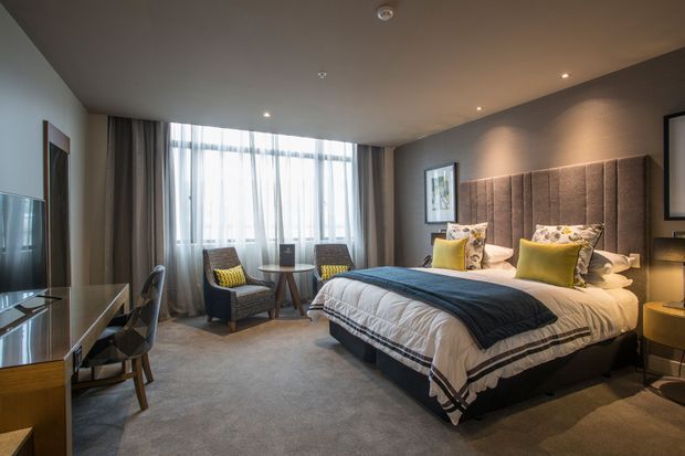 A refined contemporary bedroom with a large upholstered bed and tall padded headboard, layered white and charcoal bedding, and yellow accent cushions. Full-height windows with sheer curtains and heavier drapes provide abundant natural light. The room also includes a small sitting area, desk and chair, bedside tables, wall-mounted artwork, recessed ceiling lighting, and soft grey carpet.