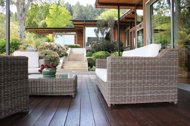 A refined outdoor lounge terrace beside a modern timber-and-glass residence. Wicker lounge chairs and a low table with white cushions sit on a dark timber deck, overlooking landscaped planting and a stepped path. The home features broad glazing, warm timber cladding and projecting flat roof planes, set within a leafy garden.