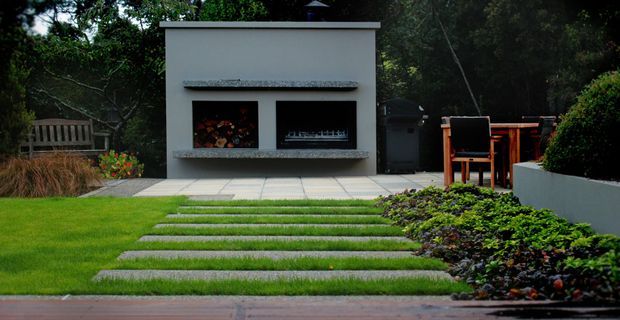 A landscaped backyard featuring a modern rendered outdoor fireplace structure with two recessed openings, one stacked with firewood and the other containing a gas fire. A tiled terrace with outdoor dining furniture sits beside clipped hedging and planting, while broad lawn and regularly spaced concrete stepping slabs lead toward the fireplace.