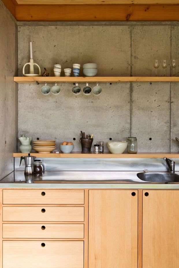 A compact, carefully detailed kitchen with exposed concrete walls, warm timber ceiling beams and open timber shelves. Light timber cabinetry with round black pulls sits below a stainless-steel benchtop and integrated sink. Ceramic dishes, cups and hanging glasses are displayed on the shelves, creating a warm utilitarian and handcrafted feel.