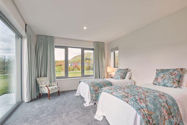 A bright, spacious twin bedroom with two single beds dressed in white bedding and blue-green floral quilts. The room has pale walls, grey carpet, full-height glazing, large windows with grey curtains, and a floral upholstered armchair. A landscaped outdoor view and hillside are visible through the windows.