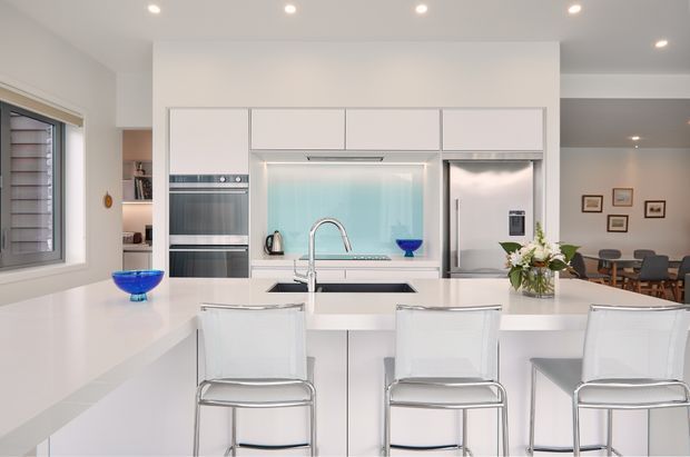 Bright contemporary white kitchen with a large waterfall-edge island and three transparent bar stools. The room features integrated stainless-steel appliances, a pale blue glass splashback, white cabinetry, recessed ceiling lights, and a dining area visible beyond the kitchen.