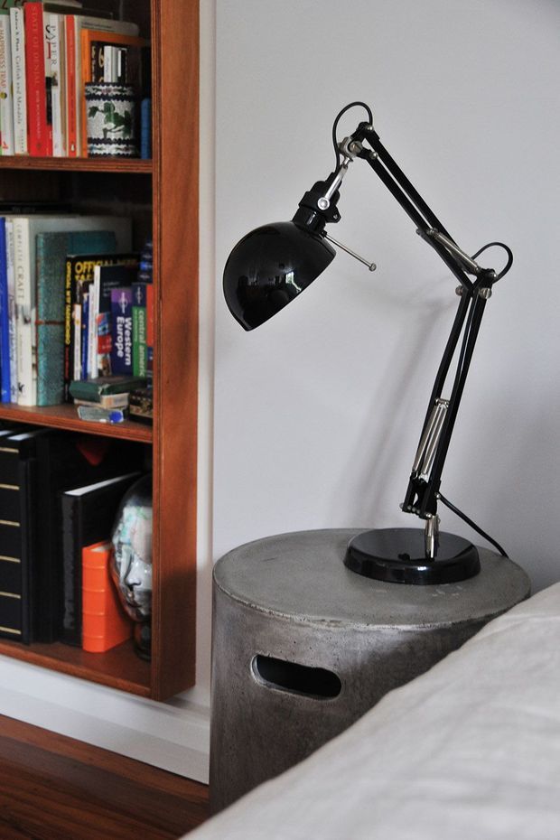Close-up interior detail of a bedroom corner, featuring a black adjustable desk lamp on a round grey concrete bedside table, beside a built-in dark timber bookshelf filled with books. A portion of the bed is visible in the foreground, with white walls and dark timber flooring creating a simple, industrial-leaning look.