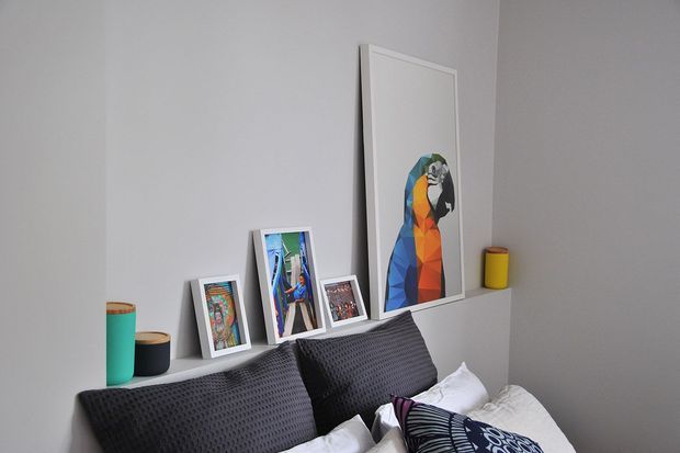 A close-up bedroom scene with a bed dressed in dark textured pillows and patterned bedding. A shallow white ledge behind the bed displays framed artwork and photographs, including a large colourful geometric parrot print, alongside two small cylindrical vessels. The room has plain light-grey walls and a restrained contemporary aesthetic.