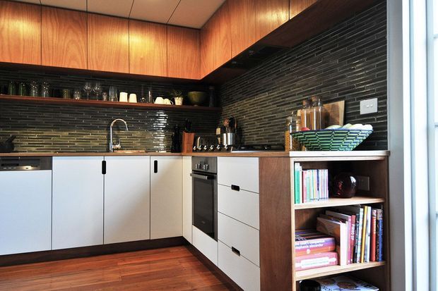 Compact L-shaped kitchen with mid-century-inspired timber overhead cabinetry, white flat-panel lower cabinets, dark horizontal brick-look splashback, stainless-steel appliances, and warm timber flooring. Open shelving displays books and kitchenware beside the cabinetry.