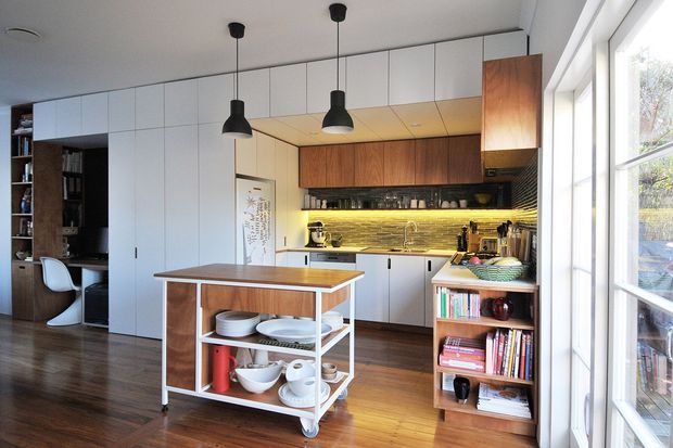 A bright, compact contemporary kitchen viewed from an adjoining living or study area. The space features extensive white flat-panel cabinetry, warm timber overhead cabinetry and shelving, a slim island or mobile kitchen cart with open storage, integrated appliances, a textured horizontal backsplash, and three black pendant lights. Large windows on the right provide abundant daylight, while dark timber flooring and built-in book storage extend into the adjacent area.